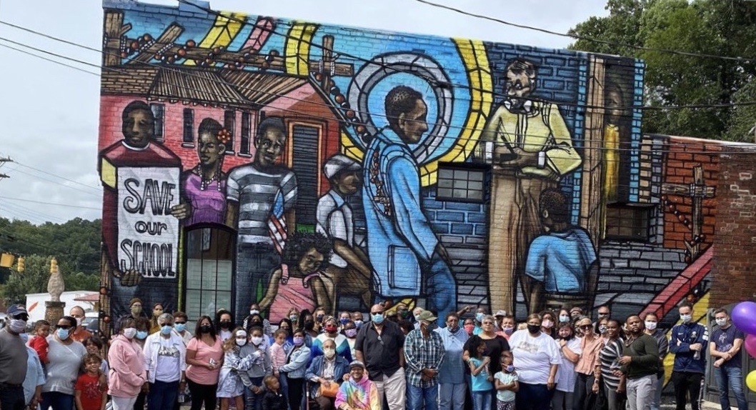 Community members gathered in front of the mural on Main Street in Old Fort, North Carolina. The mural shows Black school children carrying a “Save Our School” sign and Albert Joyner Sr. on the school steps.